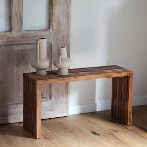 Op de afbeelding: Een houten bank met een lattenontwerp, in een warme bruine kleur. Twee beige keramische vazen met unieke vormen staan erop. De scène speelt zich af tegen een lichtgekleurde muur en een houten deur.