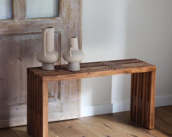Op de afbeelding: Een houten bank met een lattenontwerp, in een warme bruine kleur. Twee beige keramische vazen met unieke vormen staan erop. De scène speelt zich af tegen een lichtgekleurde muur en een houten deur.
