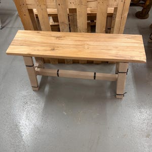 Puede incluir: Un banco rectangular de madera con una superficie superior de madera natural de color marrón claro con veta lisa. La base consta de cuatro patas verticales y una viga de soporte horizontal, construidas con madera clara de estrías verticales y unidas por bandas negras.