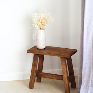 Könnte beinhalten: Ein kleiner, gebeizter Holzschemel mit rechteckiger Oberseite und abgewinkelten Beinen. Eine weiße Keramikkrug mit getrockneten Blumen steht auf dem Schemel. Die Blumen sind beige und cremefarben. Ein hellgrauer Vorhang ist im Hintergrund.