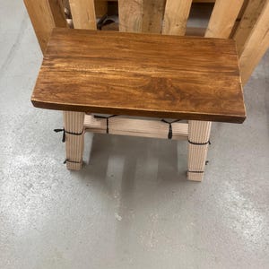 Könnte beinhalten: Ein rechteckiger Holzhocker mit einer dunkelbraun gebeizten Platte und vier hellen, vertikal gerippten Beinen. Die Beine sind mit dünnen schwarzen Bändern befestigt, die um den oberen und unteren Bereich jedes Stützpfostens gewickelt sind.