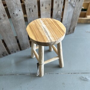 Op de afbeelding: Een ronde houten kruk met een lichtbruine natuurlijke houtnerfafwerking en een zichtbare verticale barst in het midden van de zitting. De kruk wordt ondersteund door vier cilindrische poten die verbonden zijn door een kruisvormige steunstructuur.