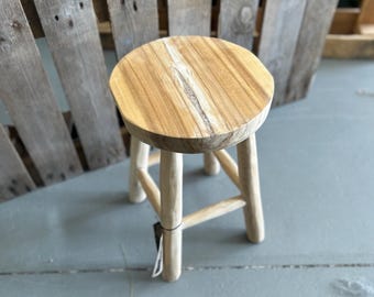 Op de afbeelding: Een ronde houten kruk met een lichtbruine natuurlijke houtnerfafwerking en een zichtbare verticale barst in het midden van de zitting. De kruk wordt ondersteund door vier cilindrische poten die verbonden zijn door een kruisvormige steunstructuur.