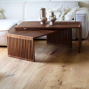 May include: Two wooden slat-style coffee tables in a warm brown hue, one partially tucked under the other. Two beige ceramic vases sit atop the larger table. The tables are on a light wood floor, with a white sofa in the background.