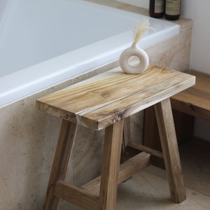 May include: A wooden stool with a natural finish and a simple design. The stool is made of solid wood and has a rectangular top. It is sitting in a bathroom next to a bathtub and a white ceramic vase with dried flowers.