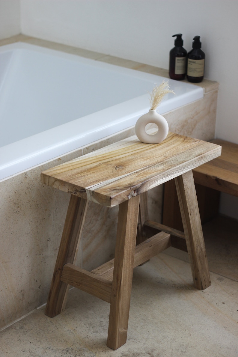 May include: A wooden stool with a natural finish and a simple design. The stool is made of solid wood and has a rectangular top. It is sitting in a bathroom next to a bathtub and a white ceramic vase with dried flowers.