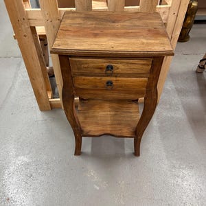 Könnte beinhalten: Ein kleiner Nachttisch aus Holz mit zwei Schubladen und einem unteren Regal. Der Nachttisch hat eine warme, braune Oberfläche und geschwungene Beine. Die Schubladen haben schwarze Ringgriffe. Der Nachttisch ist aus Holz.