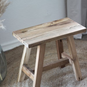 May include: A rustic wooden stool with a natural wood finish. The stool has a rectangular top and four legs. The stool is sitting on a woven rug.