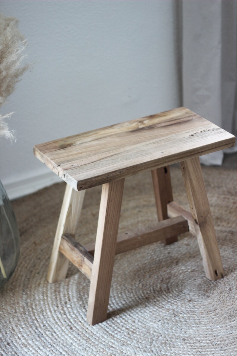 May include: A rustic wooden stool with a natural wood finish. The stool has a rectangular top and four legs. The stool is sitting on a woven rug.