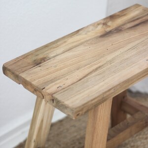 May include: A rustic wooden stool with a simple design. The stool is made of natural wood with a light brown finish. It has a rectangular top and three legs.