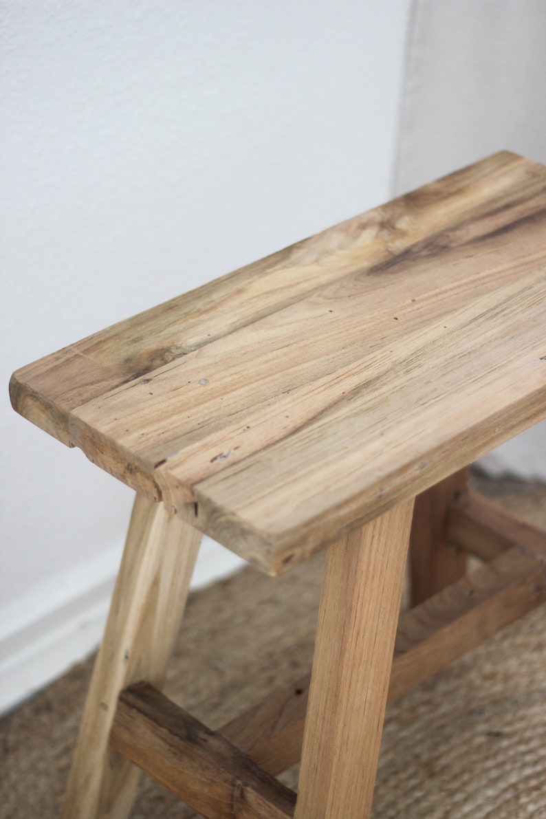 May include: A rustic wooden stool with a simple design. The stool is made of natural wood with a light brown finish. It has a rectangular top and three legs.