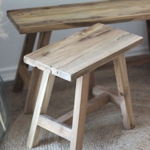 Könnte beinhalten: Ein Holzhocker mit rechteckiger Oberseite und vier Beinen. Der Hocker ist aus Naturholz mit rustikalem Finish. Der Hocker steht auf einem gewebten Teppich.