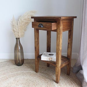 Könnte beinhalten: Ein kleiner Beistelltisch aus Holz mit einer Schublade und einem unteren Regal. Der Tisch ist aus warm getöntem Holz gefertigt und hat einen dunklen Metallgriff an der Schublade. Ein Buch liegt auf dem Regal. Eine Vase mit Trockenblumen steht daneben.