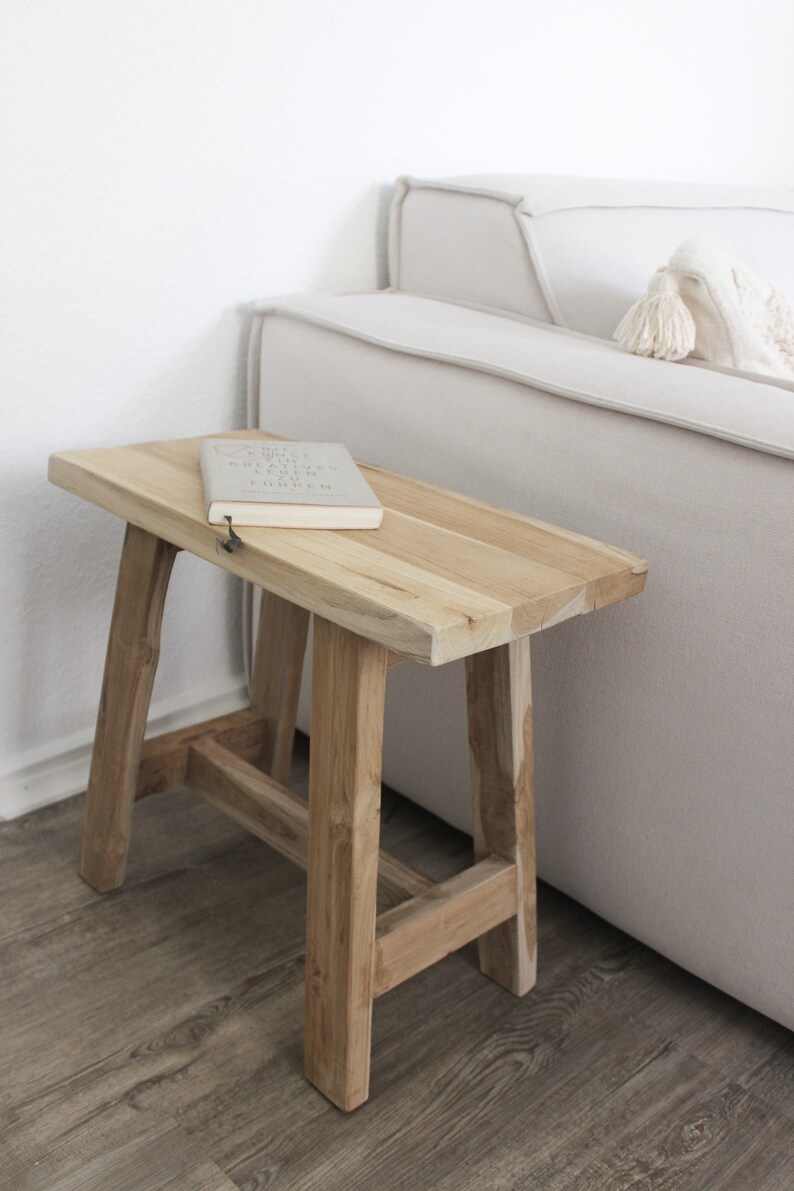 May include: A light wood side table with a book on top. The table has a simple design with four legs and a rectangular top. The book is closed and has a white cover with black text.