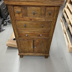 K&ouml;nnte beinhalten: Ein Holzschrank mit warmbraunem Finish. Er verf&uuml;gt &uuml;ber mehrere Schubladen und einen zweit&uuml;rigen Schrank am unteren Ende. Der Schrank hat eine rechteckige Form und wird wahrscheinlich zur Aufbewahrung verwendet. Die Beschl&auml;ge sind aus dunklem Metall.