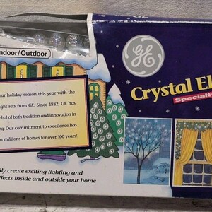 May include: A GE Crystal Elegance 70 light specialty set in a blue rectangular box. The packaging features illustrations of a house, trees, and a Christmas tree, with text stating Indoor/Outdoor and 70 Light Crystal Elegance Specialty Set.