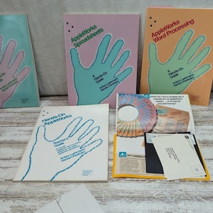 May include: A collection of vintage AppleWorks software guides and related materials. The guides feature a hand graphic and titles like "Data Bases," "Spreadsheets," and "Word Processing." Additional items include a CD-ROM, floppy disks, and other printed materials.