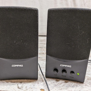May include: A pair of black Compaq computer speakers with mesh grilles. The right speaker features three circular control knobs labeled voice, volume, and power, along with a small green LED indicator light. Both units have a dark gray base.
