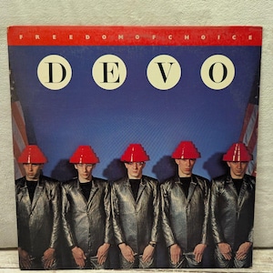 May include: A vinyl record cover for Devo's Freedom of Choice featuring five individuals in metallic silver suits and red tiered hats against a blue background with American flag elements on the sides and the text DEVO in white circles.