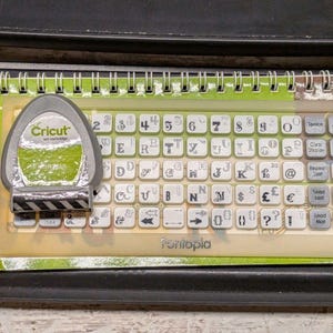 May include: A Cricut Fontopia art cartridge and a corresponding plastic keyboard overlay. The overlay features a grid of white square keys with black letters, numbers, symbols, and command labels like Space, Back Space, and Load Mat printed on them.