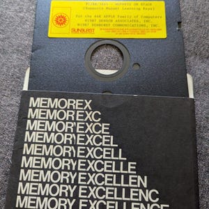 May include: A vintage black 5.25-inch floppy disk with a yellow label that reads "MUPPETS ON STAGE." The disk is in a black sleeve with the word "MEMOREX" and variations of "MEMORY EXCELLENCE" printed in white.