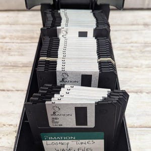 May include: A black storage case filled with numerous 3.5-inch floppy disks. The disks are black with silver labels, some bearing the "IMATION" brand. A handwritten label reads "Looney Tunes WAVE. Files THEME."