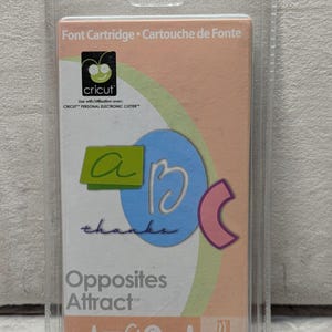 May include: A Cricut font cartridge in a clear plastic package. The cartridge features the text "Opposites Attract" and includes the letters "a", "b", and "c" in various styles. The package also displays the Cricut logo and the words "Font Cartridge".