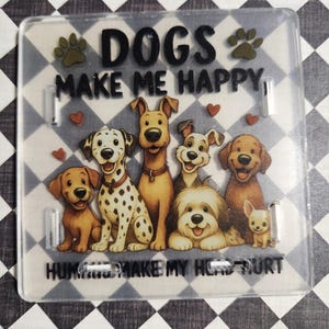 May include: A clear, rectangular acrylic coaster featuring a cartoon illustration of seven different dog breeds. The text "DOGS MAKE ME HAPPY" and "HUMANS MAKE MY HEAD HURT" is printed on the coaster, along with paw prints and hearts.