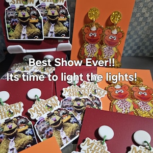 May include: A collection of Muppet Show themed earrings and display cards. The earrings feature Fozzie Bear designs with glitter accents. The display cards have the text "Best Show Ever!! Its time to light the lights!" and Muppet Show logos.