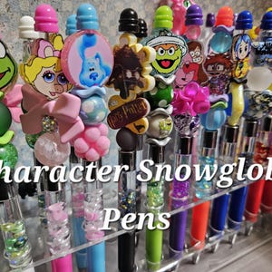 May include: A collection of character snowglobe pens with various designs. The pens feature colorful barrels and are topped with character figures, including a frog, a cartoon character with a pink bow, and Blue from Blue's Clues. The text "Character Snowglobe Pens" is visible.