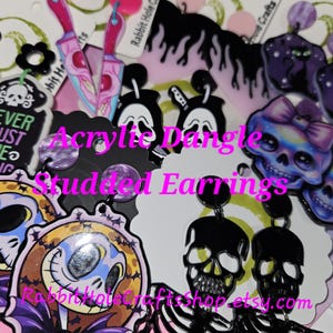 May include: A collection of acrylic dangle and studded earrings in various designs. The earrings feature a variety of spooky and gothic themes, including skulls, skeletons, and bats. The color palette includes black, pink, purple, and white. The text "Acrylic Dangle Studded Earrings" is visible.