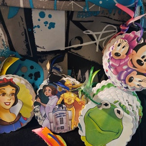 May include: A collection of colorful, round, paper crafts featuring various Disney characters. The characters include Snow White, Princess Leia, C-3PO, R2-D2, Kermit the Frog, and Mickey Mouse. Each craft is decorated with a ribbon and a bow.