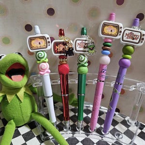 May include: A collection of decorative pens featuring designs inspired by The Muppet Show. Each pen has a unique color scheme, with details like beads, charms, and TV-shaped toppers. The pens are displayed in a clear acrylic holder, with a Kermit the Frog plushie nearby.