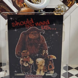 May include: A clear acrylic display case featuring a black graphic print from the movie Labyrinth. The print includes the text "Should you need us..." and images of characters from the film. Decorative elements include a crescent moon and spherical ornaments.