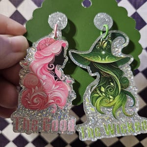 May include: A pair of glittery acrylic dangle earrings featuring silhouettes of two figures. One earring shows a pink-haired figure with the text The Good, and the other shows a green-haired figure in a hat with the text The Wicked.