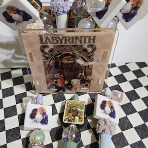 May include: A collection of pens and decorative items featuring imagery from the movie "Labyrinth." The pens have glittery barrels and decorative tops. The display box has the movie title and characters. The background is a black and white checkered pattern.