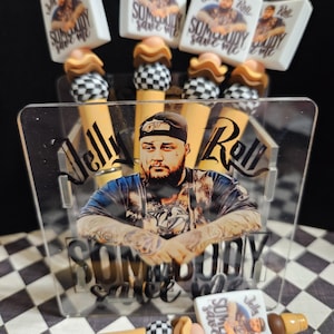 May include: A collection of decorative pen-shaped items featuring the text "Jelly Roll" and "Somebody Save Me". The pens have a variety of designs, including a checkered pattern, a portrait, and a light brown and tan color scheme.