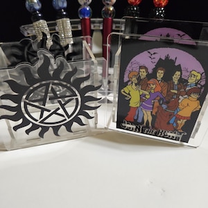 May include: A collection of pens and acrylic display stands. The pens have decorative tops with images and beads. One stand features a black and white pentagram design, and another displays a cartoon image with the text "Join the Hunt".