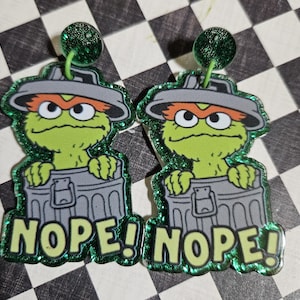 May include: A pair of novelty earrings featuring a green cartoon character in a trash can, with the word "NOPE!" in green letters. The earrings have a glittery green border and a green stud.