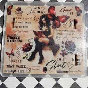 May include: A clear, rectangular plastic card with a printed design featuring a woman reading a book, butterflies, roses, and text. The text includes phrases like "Talk Dirty to Me" and "Smut Slut."