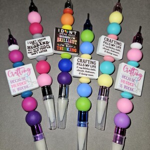 May include: A collection of colorful lip gloss pens with beaded accents. Each pen features a unique combination of beads in various colors, and a small rectangular charm with a humorous quote about crafting. The pens are designed for cosmetic use.
