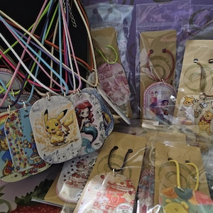 May include: A collection of colorful pendant necklaces featuring cartoon characters like Pikachu, Ariel, and Winnie the Pooh. The pendants are on various colored cords and packaged in clear plastic. The background is a mix of purple and red.