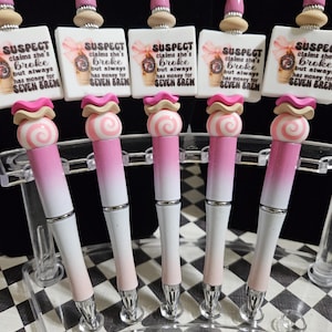 May include: A collection of five decorative pens with a pink and white gradient design. Each pen features a decorative top with a pink and white swirl design and a small square with the text "SUSPECT claims she's broke but always has money for SEVEN BREM."