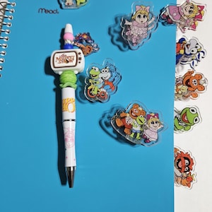 May include: A blue spiral-bound notebook with a pen and several character-shaped magnets. The pen has a white barrel with pink accents and a TV-shaped top. The magnets feature various Muppet Show characters.