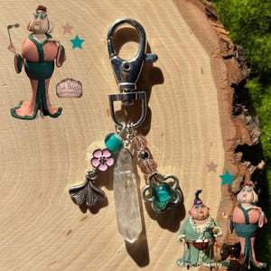 May include: Silver-tone keychain with a lobster clasp, adorned with a clear crystal point, pink flower charm, and a ginkgo leaf charm. Additional charms include pink beads and a teal-colored bead. Decorative figurines are also present.