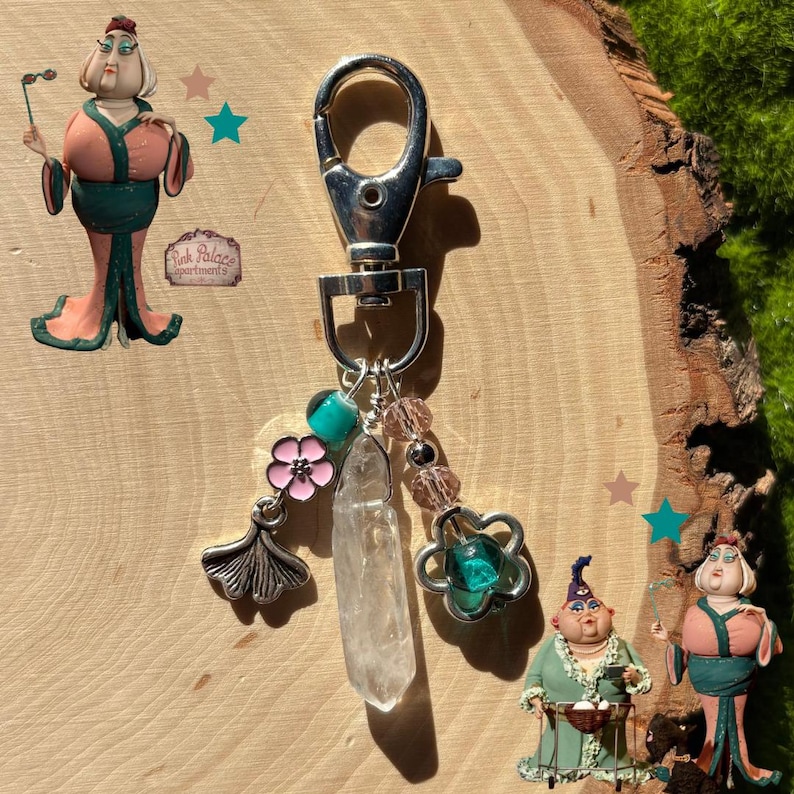 May include: Silver-tone keychain with a lobster clasp, adorned with a clear crystal point, pink flower charm, and a ginkgo leaf charm. Additional charms include pink beads and a teal-colored bead. Decorative figurines are also present.