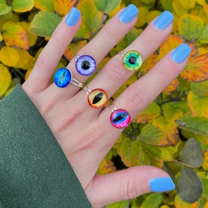 May include: Five silver rings with colorful eye-shaped glass stones. The rings are worn on a hand with blue nail polish.