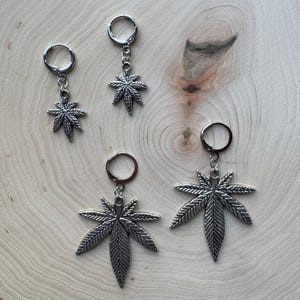 May include: Three pairs of silver earrings with a marijuana leaf charm. The earrings are on a wooden surface.