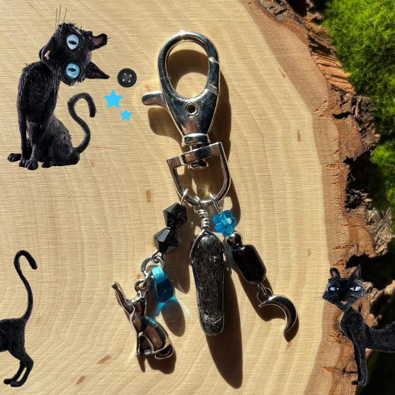 May include: A silver-toned keychain with black and blue beads, a small cat charm, and a black stone pendant. The keychain has a lobster clasp and is decorated with a crescent moon charm. The background features cartoon cat illustrations.