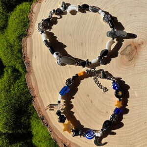 K&ouml;nnte beinhalten: Zwei Armb&auml;nder mit Perlen mit himmlischen und abstrakten Designs. Ein Armband hat schwarze, wei&szlig;e und graue Perlen mit Mond- und Sternanh&auml;ngern. Das andere enth&auml;lt blaue, gelbe und schwarze Perlen mit Stern- und Mondanh&auml;ngern.